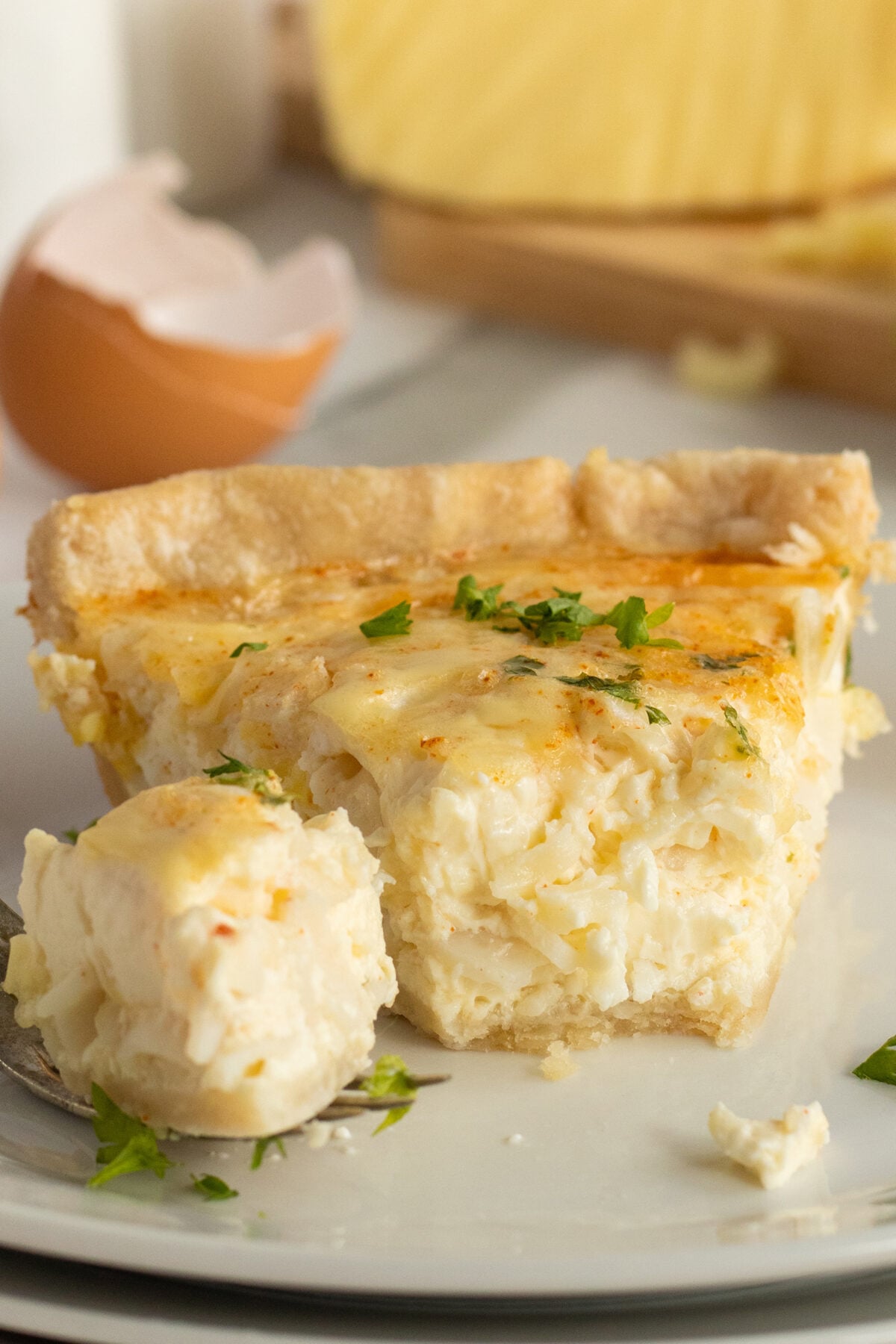 A close-up of a slice of cheesy quiche on a plate, garnished with parsley. A fork holds a bite-sized piece in the foreground, and an eggshell is visible in the background.