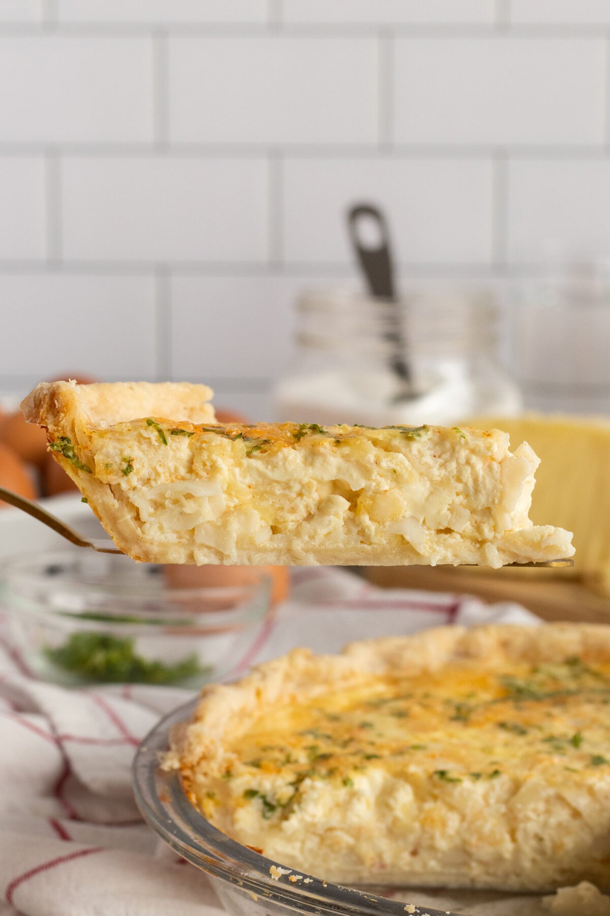 A close-up of a slice of quiche being lifted from a pie dish, showing its creamy filling and flaky crust. In the background, ingredients like eggs and cheese are visible on a kitchen counter.