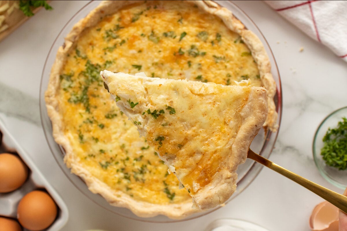 A close-up of a slice of quiche with a golden, flaky crust and sprinkled herbs being lifted from a whole quiche in a glass pie dish. Eggs and chopped herbs are visible nearby on a white surface.