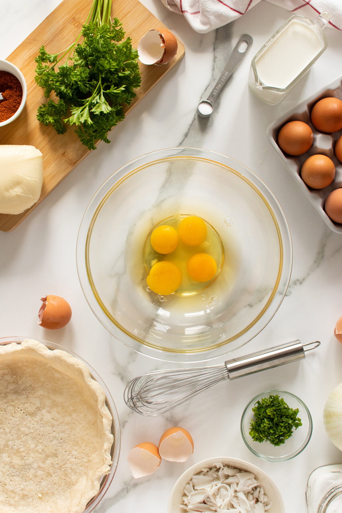 A glass bowl with three cracked eggs is on a marble surface, surrounded by a pie crust, whisk, fresh parsley, cheese, cream, brown eggs, broken eggshells, seasonings, and onions, ready for baking.