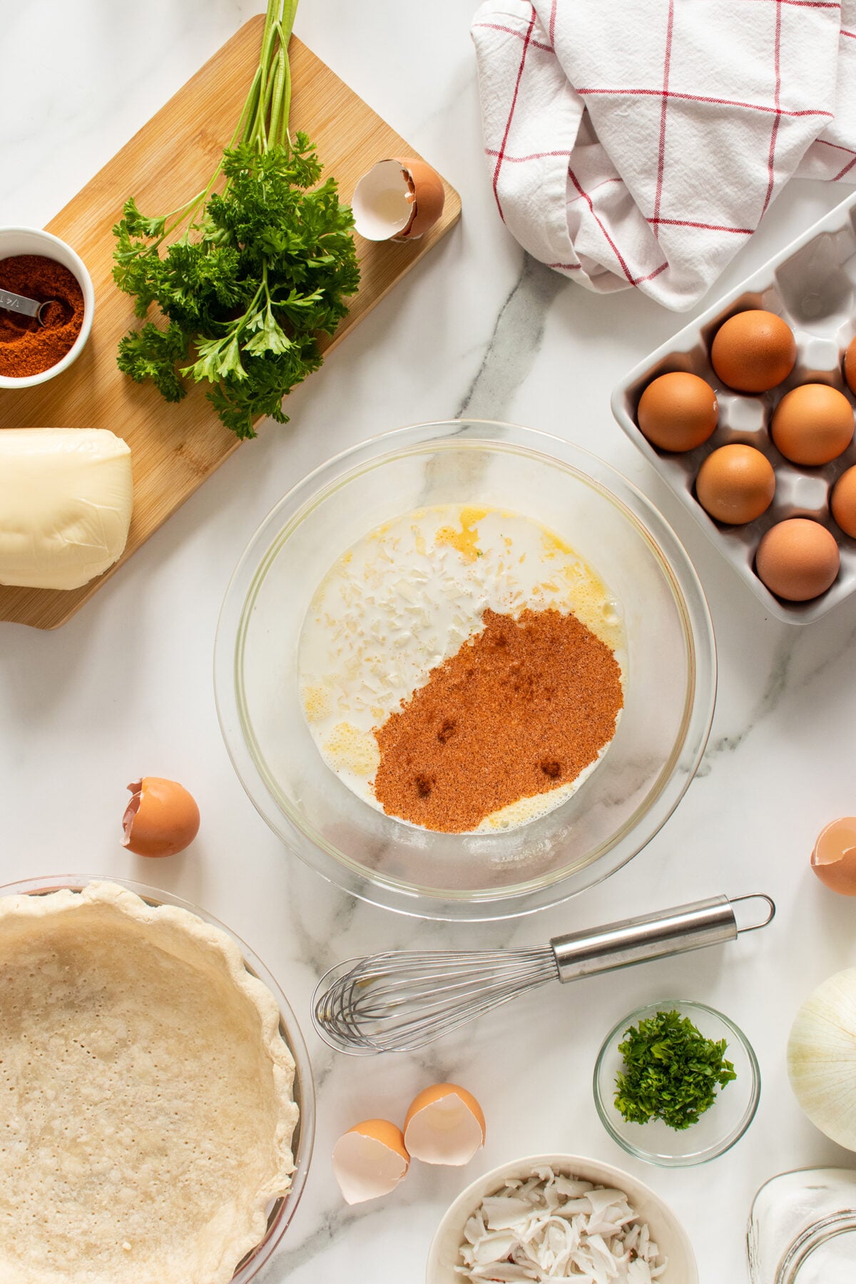 Top-down view of baking ingredients on a marble surface, including eggs, spices, parsley, cheese, a pie crust, and a mixing bowl with a whisk, surrounded by cracked eggshells and kitchen towels.