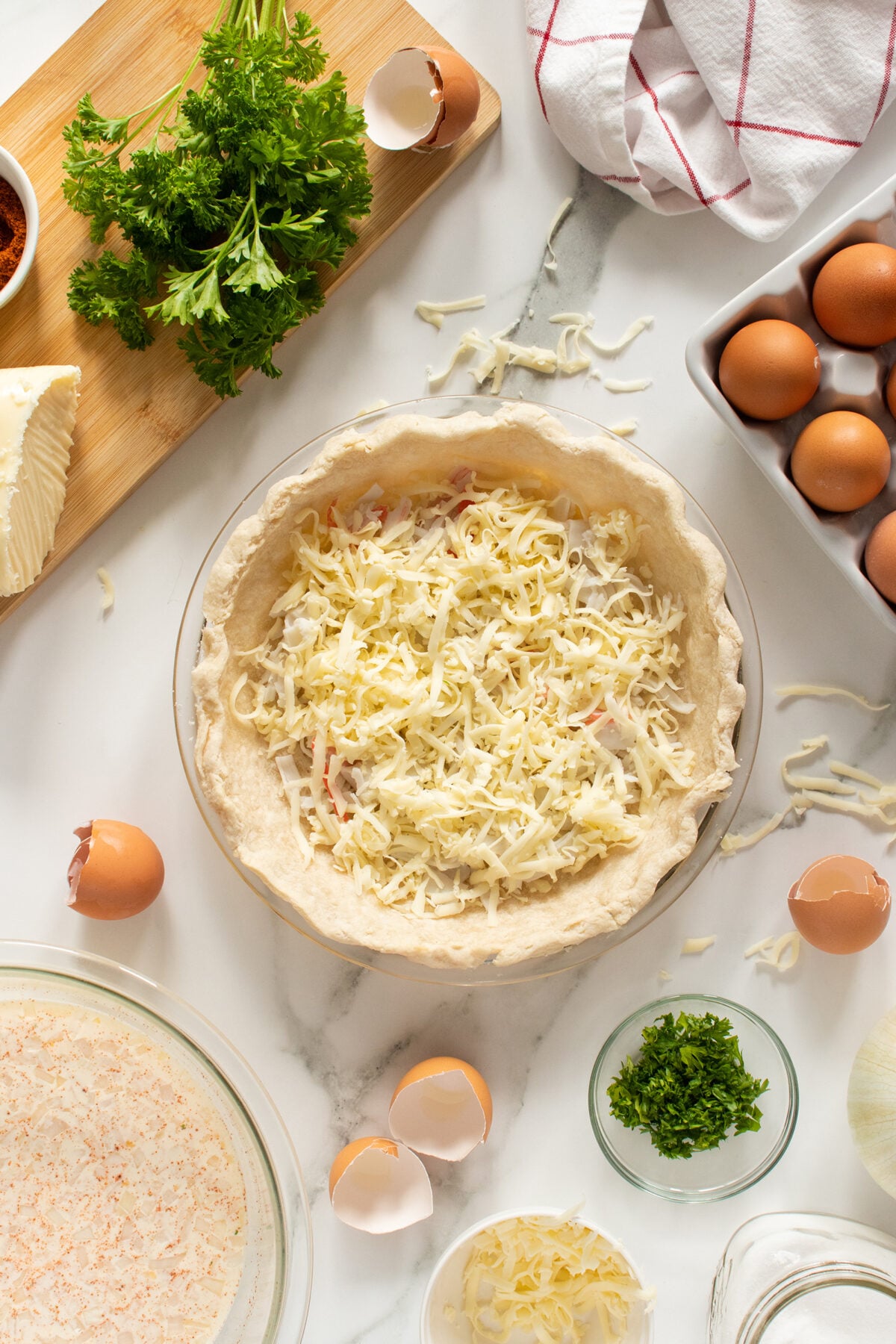 A pie crust in a glass dish filled with shredded cheese sits on a white surface surrounded by eggs, broken eggshells, chopped parsley, a cheese wedge, and a bowl of creamy mixture, suggesting a quiche preparation.