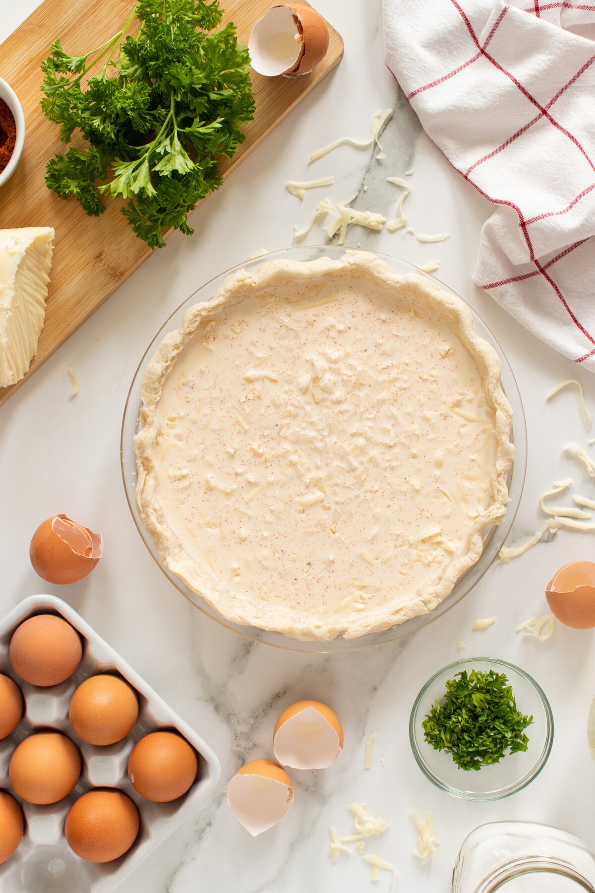 A pie crust filled with a creamy mixture sits on a marble surface surrounded by eggs, broken eggshells, shredded cheese, fresh parsley, a kitchen towel, and a wooden cutting board with parsley and cheese.