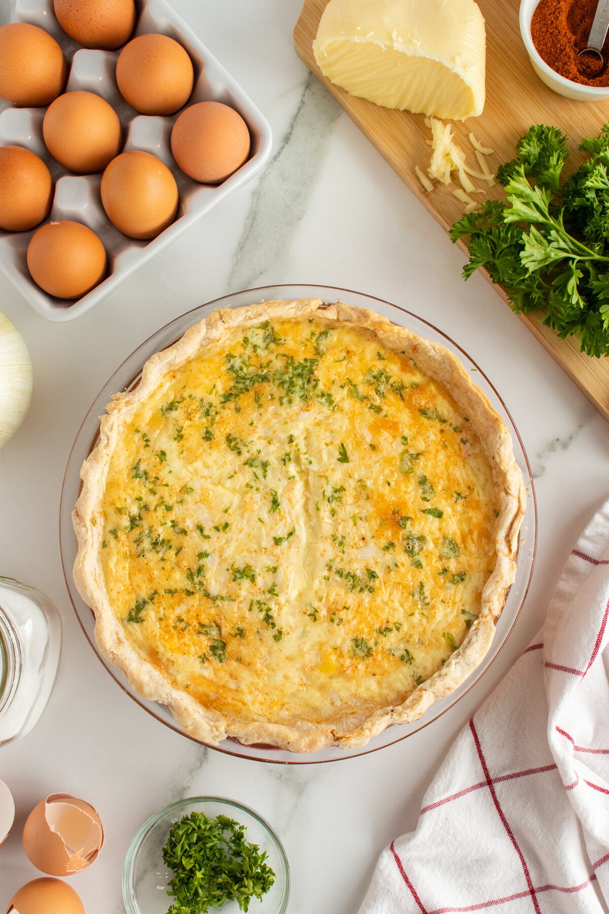 A baked quiche with a golden crust topped with chopped parsley sits on a marble countertop surrounded by whole eggs, fresh parsley, cheese, a cutting board, and a red-striped kitchen towel.
