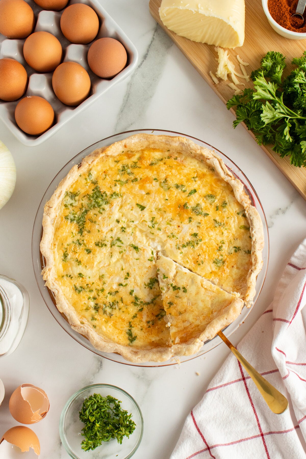 A baked quiche with a slice cut out, topped with chopped parsley, sits on a marble countertop surrounded by fresh eggs, cheese, parsley, cracked eggshells, and a kitchen towel.