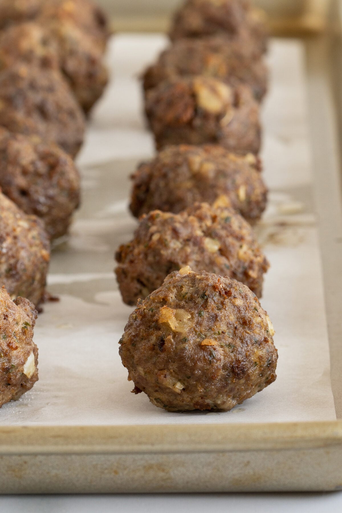 Rows of browned meatballs with visible pieces of onion on a parchment-lined baking sheet. The meatballs are evenly spaced and in focus at the front, becoming blurrier toward the back.