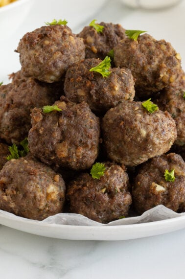 A plate stacked with browned meatballs, garnished with small sprigs of fresh parsley, sits on a white surface.