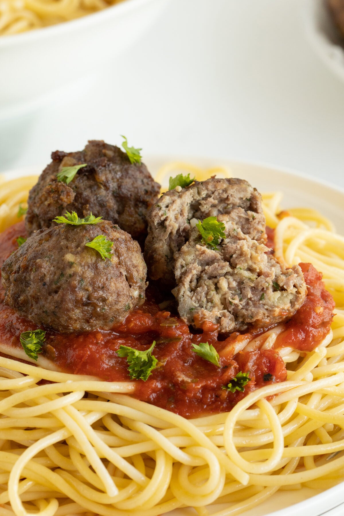 A plate of spaghetti topped with tomato sauce and three meatballs, one of which is cut open to show the inside. Chopped parsley is sprinkled on top.