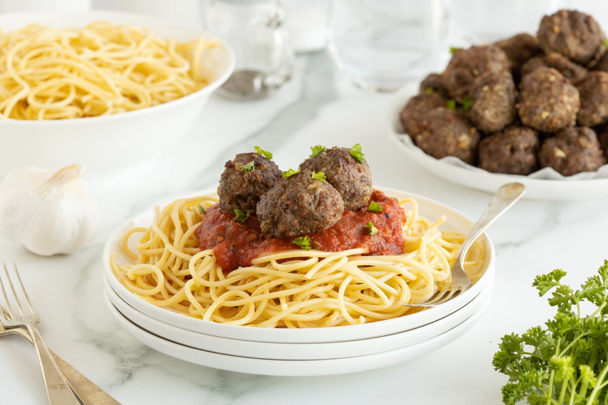 A plate of spaghetti topped with tomato sauce and three meatballs, garnished with chopped parsley. In the background, there is a bowl of plain spaghetti, a plate of meatballs, garlic, parsley, and a glass of water.