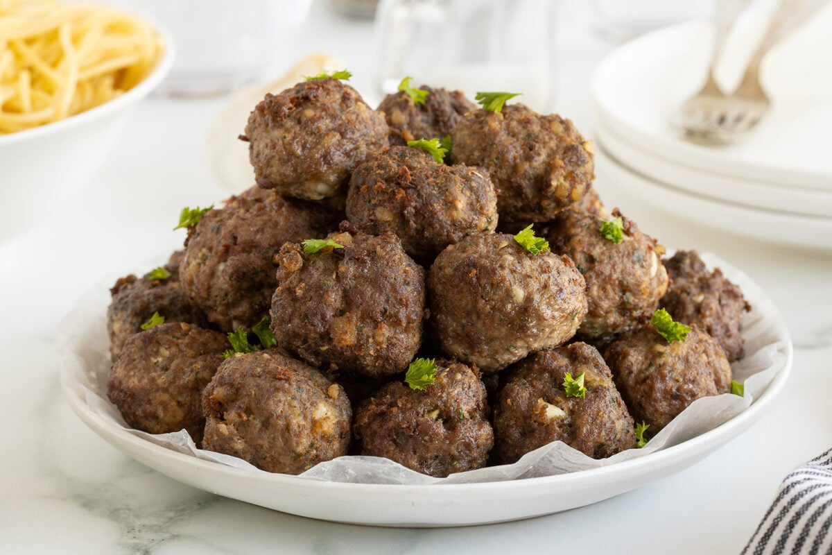 A white plate piled high with cooked meatballs garnished with fresh parsley, set on a light surface with dishes, a fork, and a bowl of spaghetti in the background.