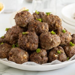 A white plate piled high with cooked meatballs garnished with fresh parsley, set on a light surface with dishes, a fork, and a bowl of spaghetti in the background.