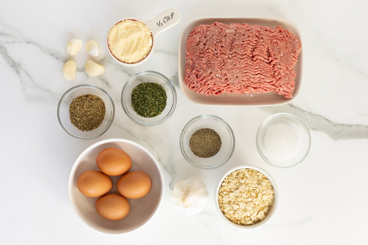 Overhead view of raw ground meat, four eggs, garlic cloves, seasonings, rolled oats, breadcrumbs, and sugar in bowls on a white marble surface.