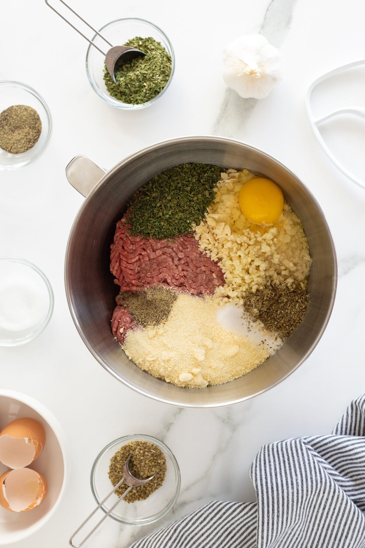 A metal mixing bowl with ground meat, an egg, minced garlic, breadcrumbs, dried herbs, and spices, surrounded by small bowls of seasonings and eggshells on a white countertop.