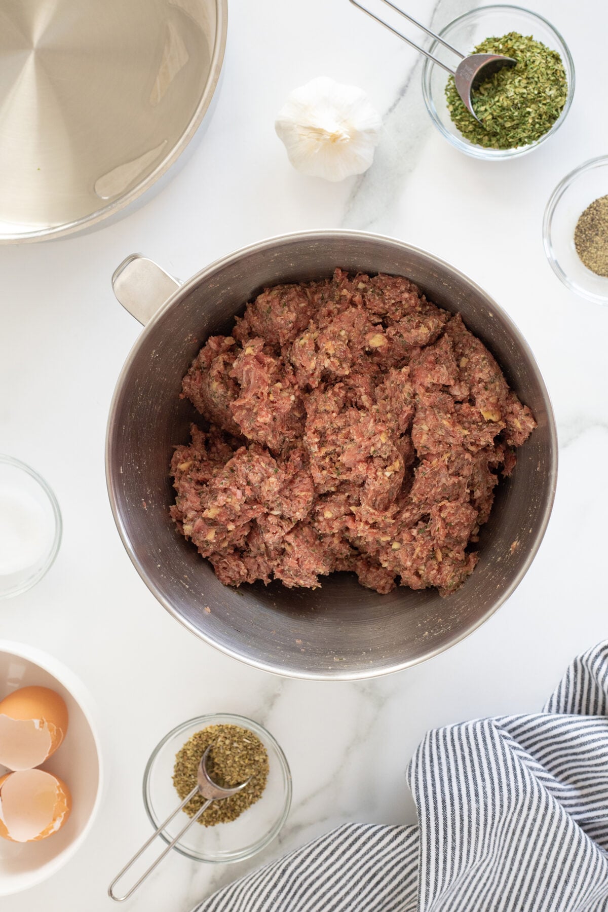 A metal bowl filled with ground meat mixture sits on a marble countertop, surrounded by spices in small bowls, cracked eggshells, a head of garlic, and a striped kitchen towel.