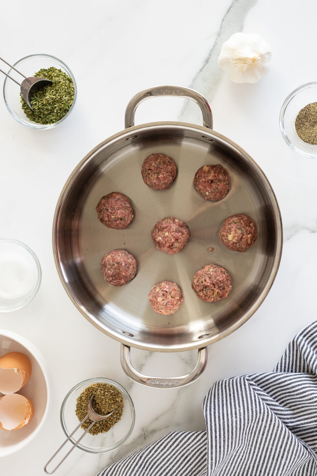 A metal pan with seven raw meatballs sits on a marble surface, surrounded by bowls of herbs, cracked eggshells, a whole garlic bulb, and a striped cloth.