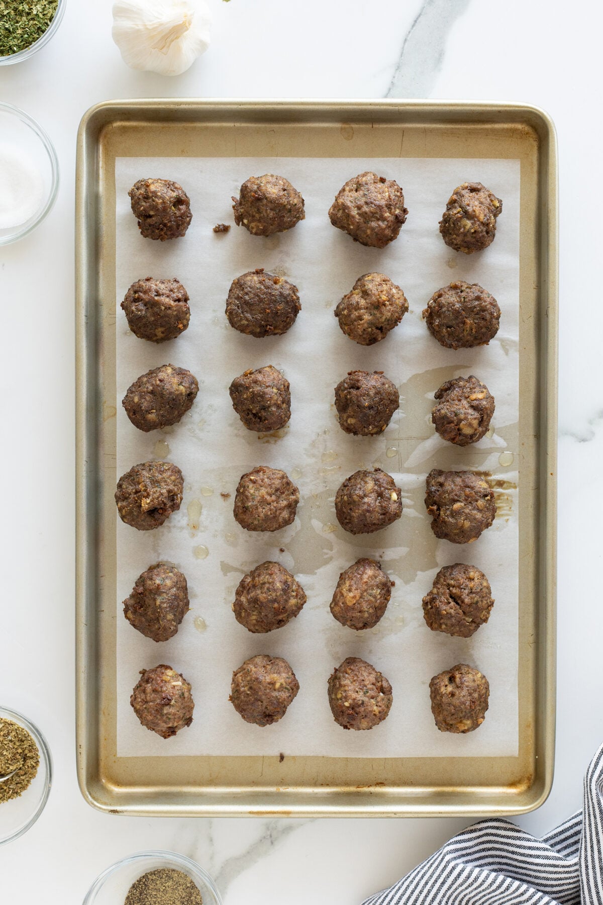 A baking sheet lined with parchment paper holds 24 cooked meatballs arranged in rows. Surrounding the tray are small bowls of spices and a clove of garlic, all on a white marble surface.