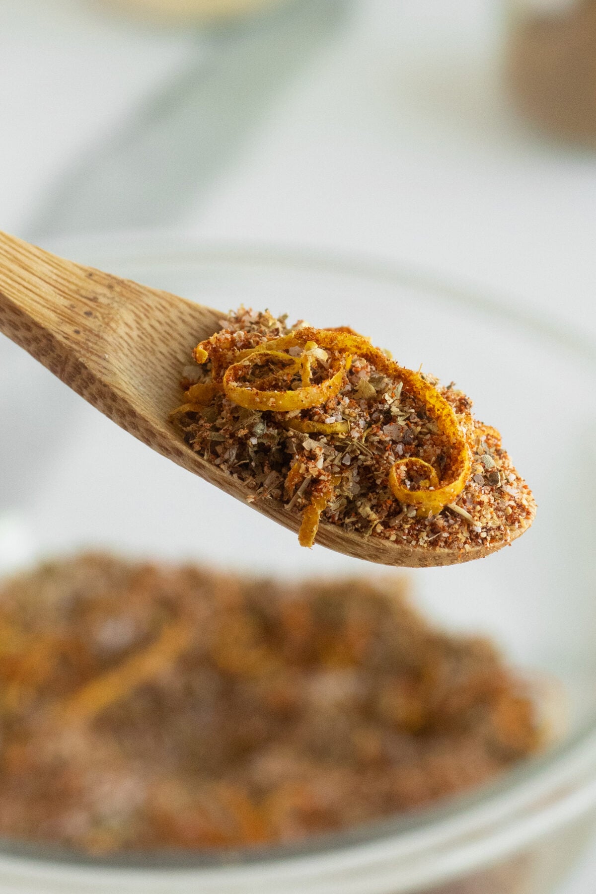 A close-up of a wooden spoon holding a blend of dry spices and orange zest, with more of the spice mixture blurred in a glass bowl in the background.