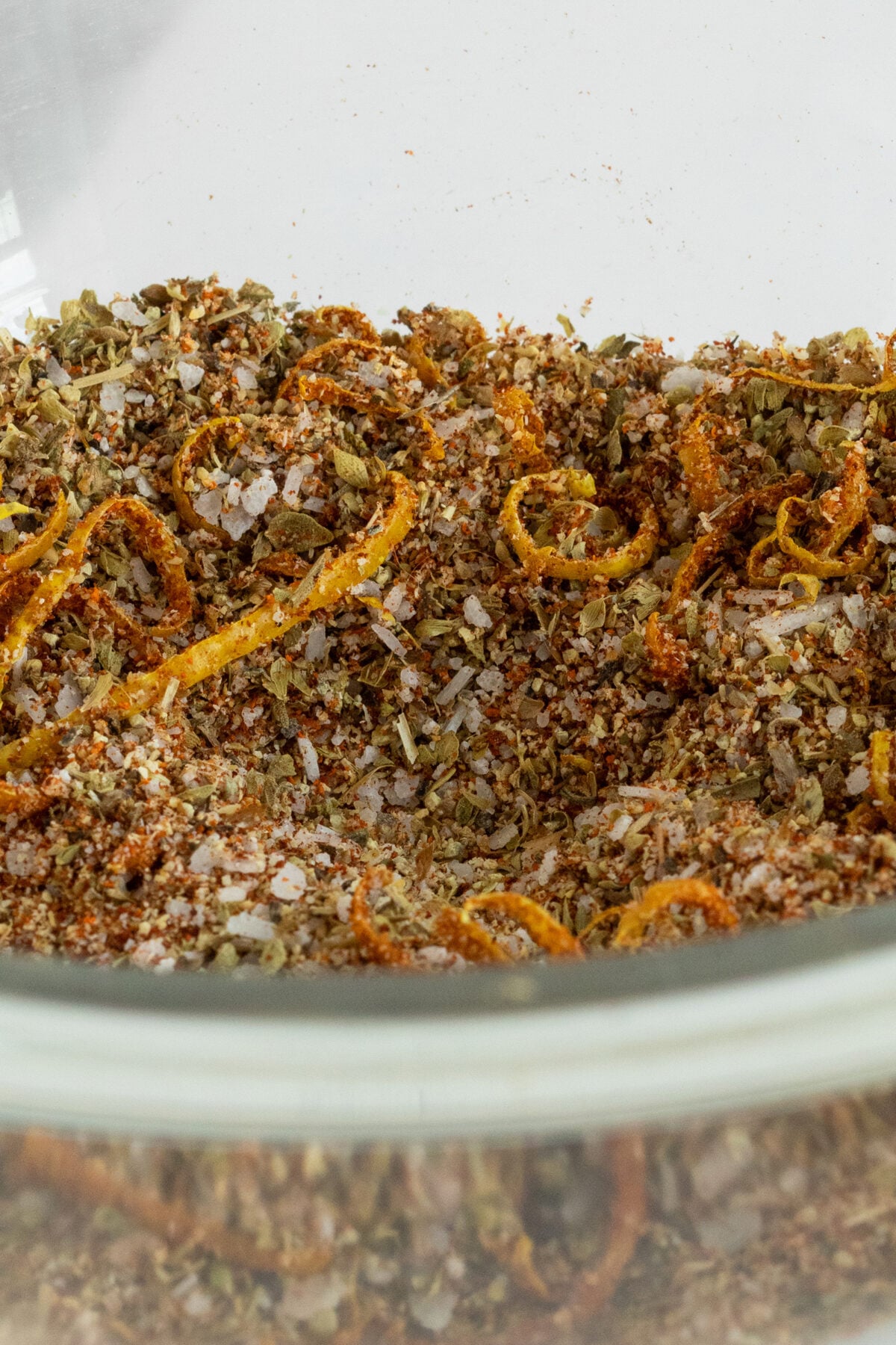 A close-up of a glass bowl containing a seasoning mix with dried herbs, coarse salt, spices, and orange peel strips.