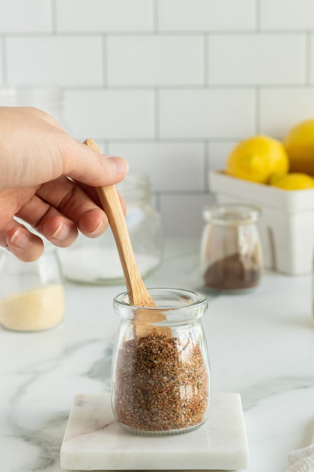A hand holds a small wooden spoon scooping ground flaxseed from a glass jar on a marble surface, with jars and lemons blurred in the background.