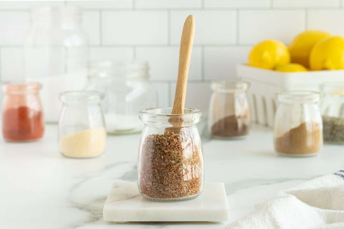 A small glass jar filled with a brown spice blend sits on a marble coaster, with a wooden spoon inside. Out-of-focus jars and a basket of lemons are in the background on a white kitchen countertop.