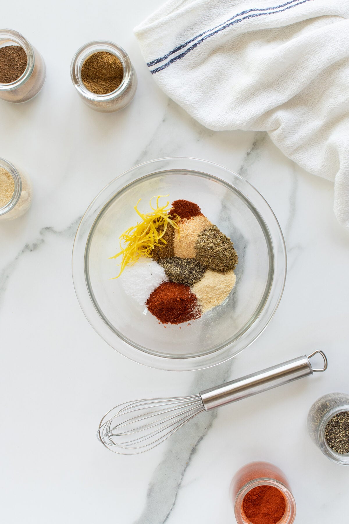 A glass bowl on a marble surface contains various spices and lemon zest, with a small whisk beside it. Spice jars, some open, and a white kitchen towel are arranged around the bowl.