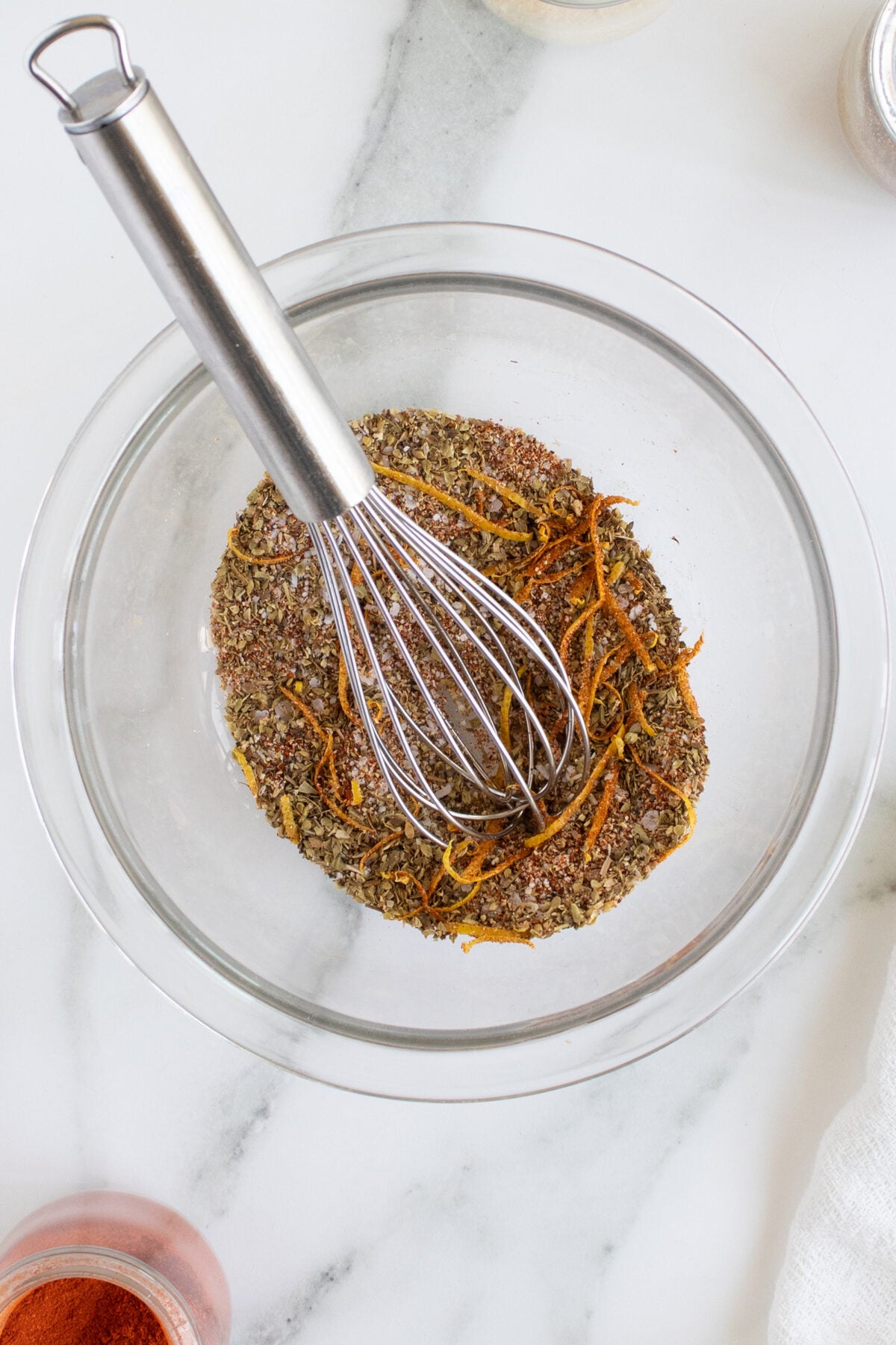 A metal whisk rests in a glass bowl containing mixed dried herbs and orange zest on a marble countertop. Nearby is a small bowl with red spice and part of a white cloth.
