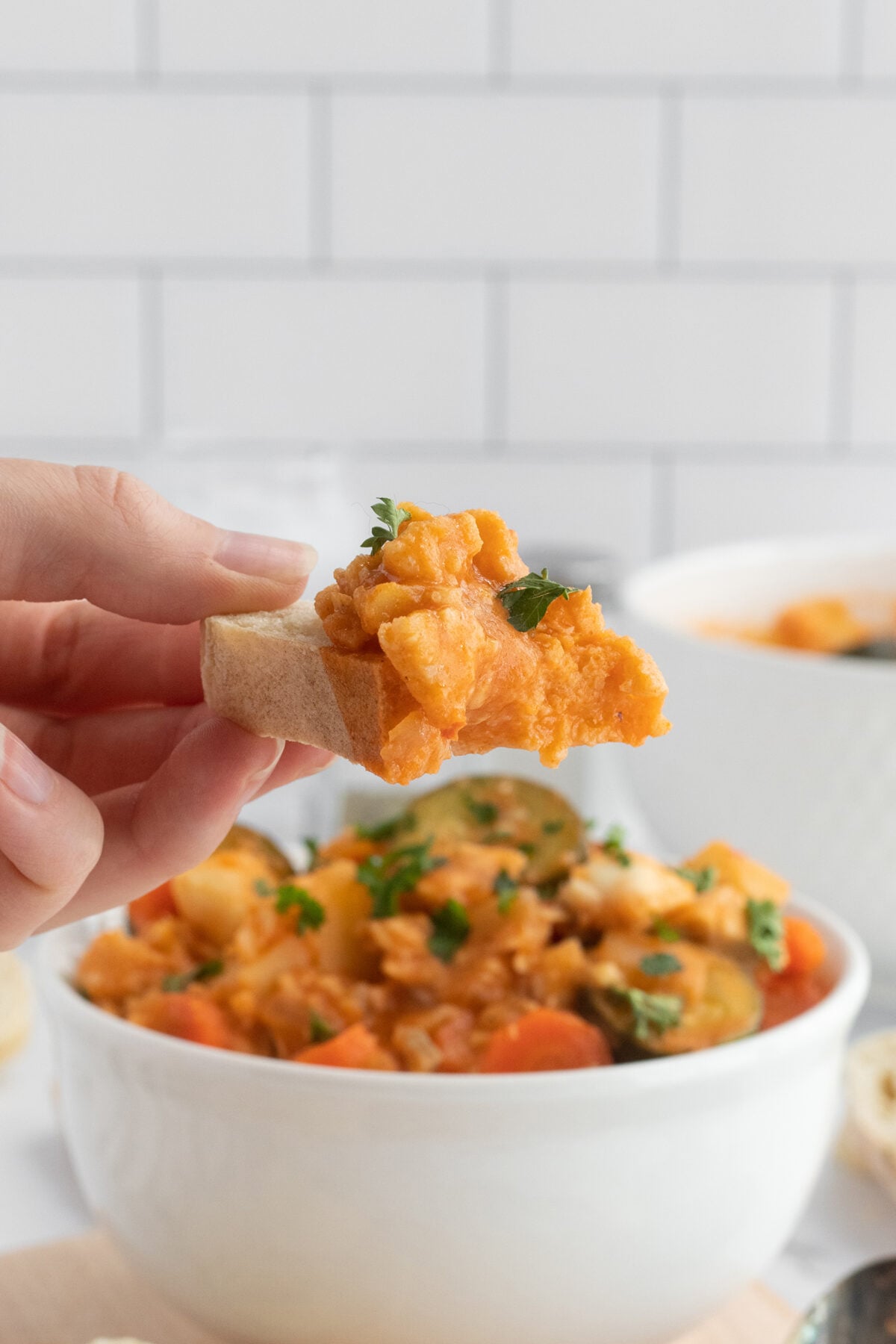 A hand holds a piece of bread topped with a chunky, orange-colored vegetable stew, garnished with parsley. In the background, a white bowl filled with the same stew is visible.