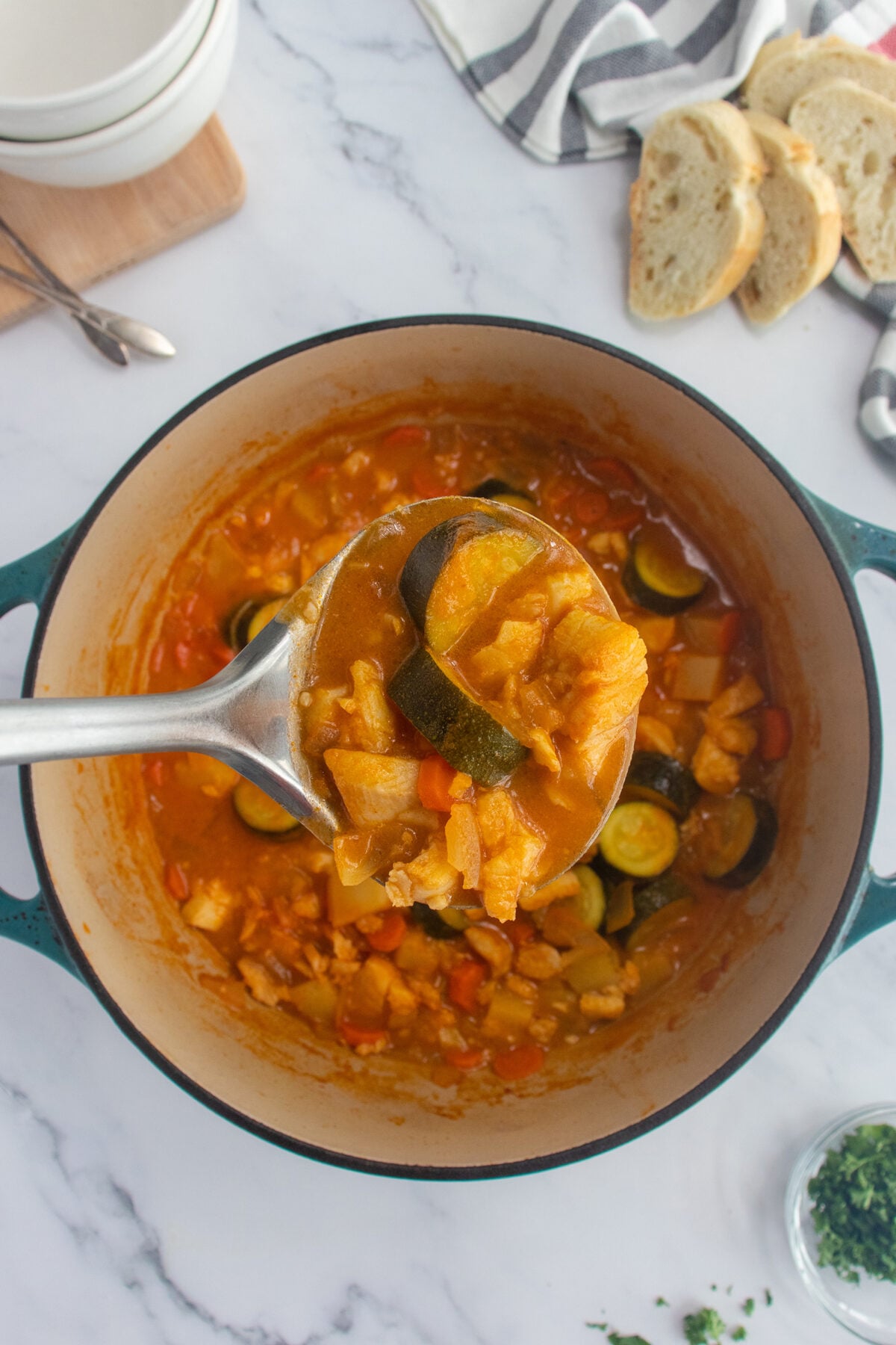 A ladle lifts a serving of chunky vegetable stew with zucchini, tomatoes, and other vegetables from a large pot. Sliced bread, bowls, and a striped towel are visible on the marble countertop nearby.