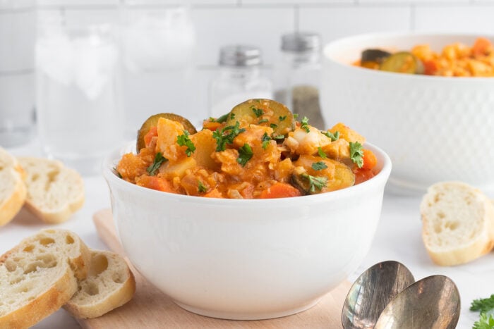 A white bowl filled with vegetable stew, garnished with parsley, sits on a wooden board surrounded by sliced baguette. Another bowl of stew and glasses of ice water are in the background.