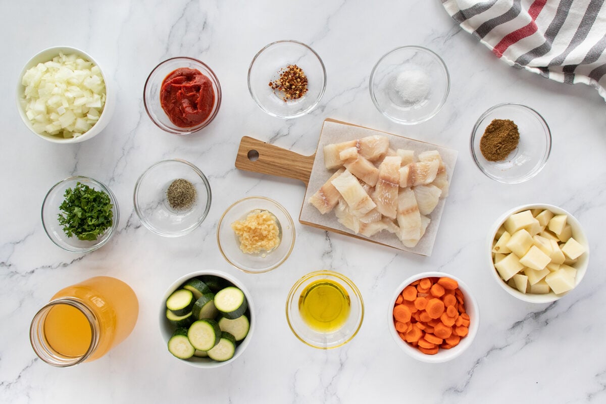 Top view of various ingredients in bowls on a marble surface, including chopped onions, tomato paste, red pepper flakes, spices, parsley, garlic, zucchini, carrots, potatoes, fish pieces, olive oil, and a jar of broth.
