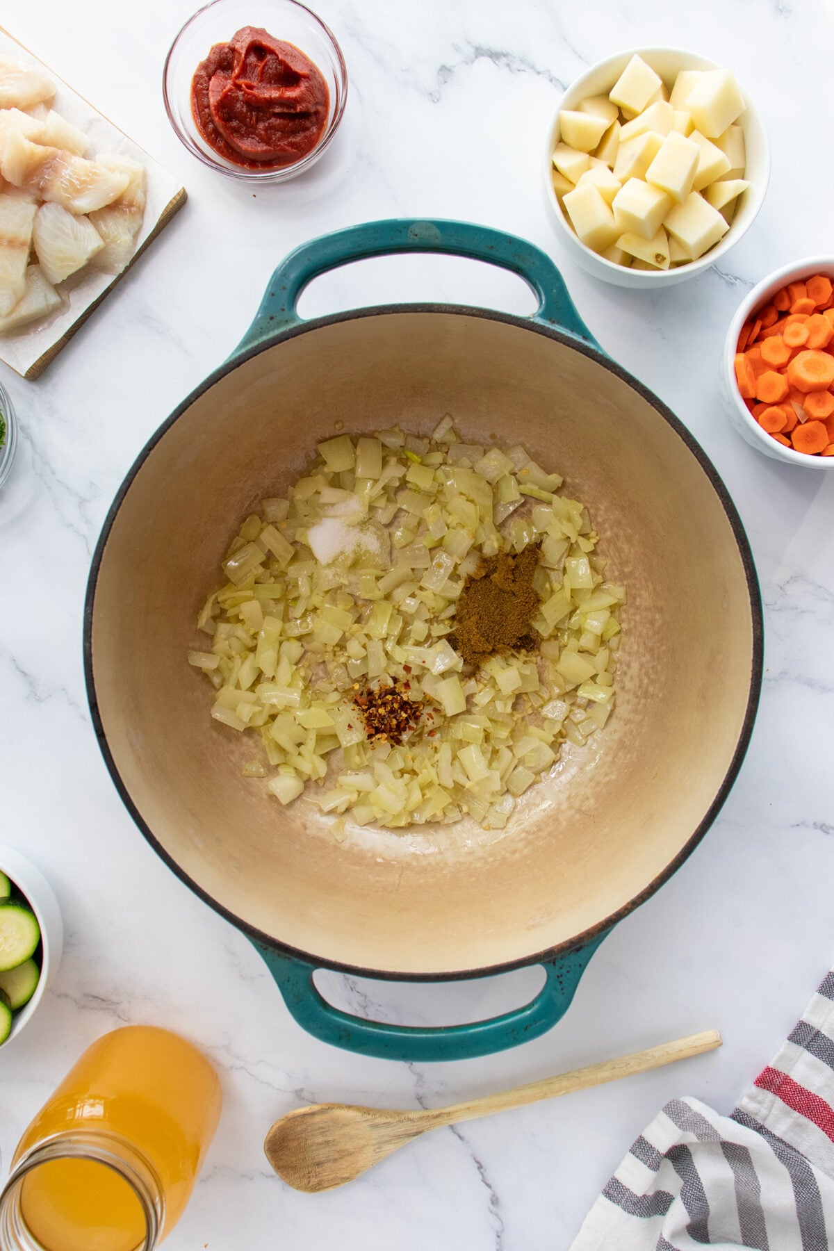 A large pot contains sautéed onions, salt, curry powder, and chili flakes. Surrounding the pot are bowls of chopped potatoes, carrots, zucchini, fish, tomato paste, and a jar of broth on a white countertop.
