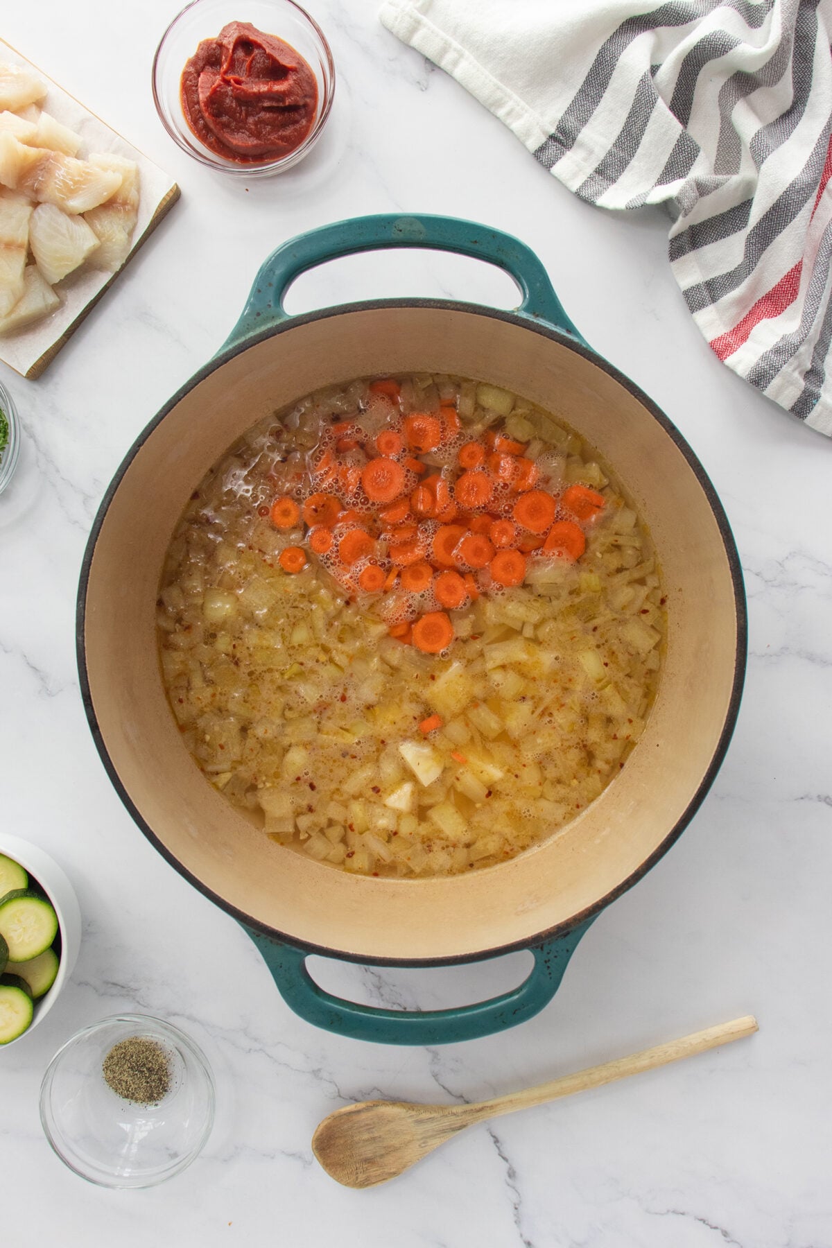 Large pot with sautéed onions and sliced carrots cooking in broth, surrounded by bowls of fish, tomato paste, spices, and zucchini on a marble countertop with a striped towel and wooden spoon nearby.