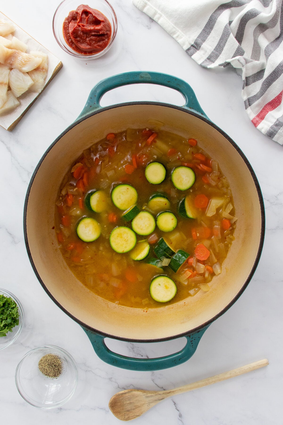 A large pot of vegetable soup with zucchini and carrots sits on a marble countertop, surrounded by chopped fish, tomato paste, spices, parsley, and a wooden spoon. A striped towel is in the background.