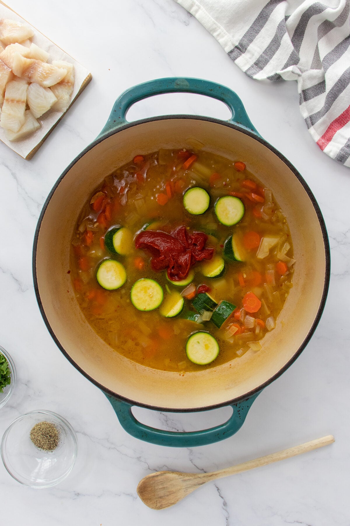 A large pot sits on a white marble surface filled with broth, sliced carrots, zucchini, onions, and a dollop of tomato paste. Nearby are a wooden spoon, spice bowls, fish pieces, and a striped towel.