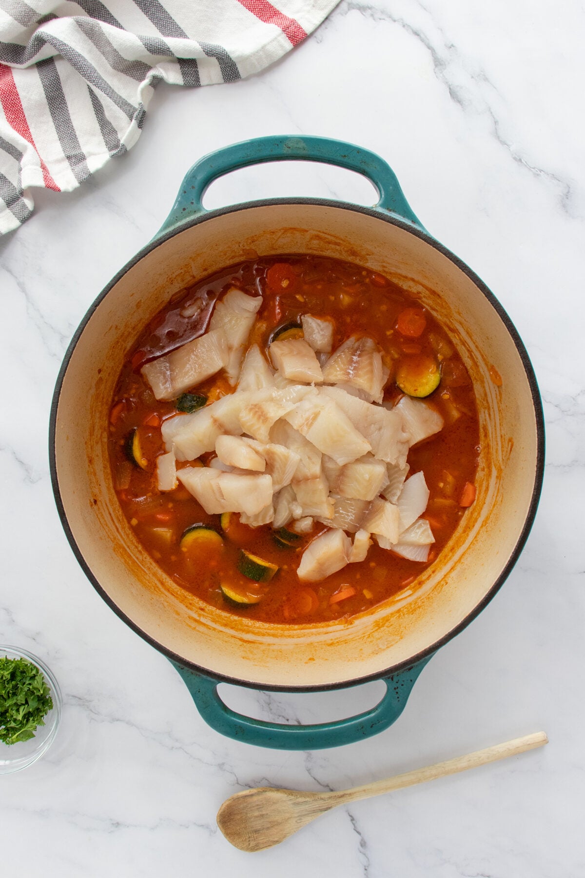 A teal Dutch oven filled with tomato-based stew, zucchini, and raw white fish pieces sits on a marble countertop. A wooden spoon, a bowl of chopped herbs, and a striped towel are nearby.