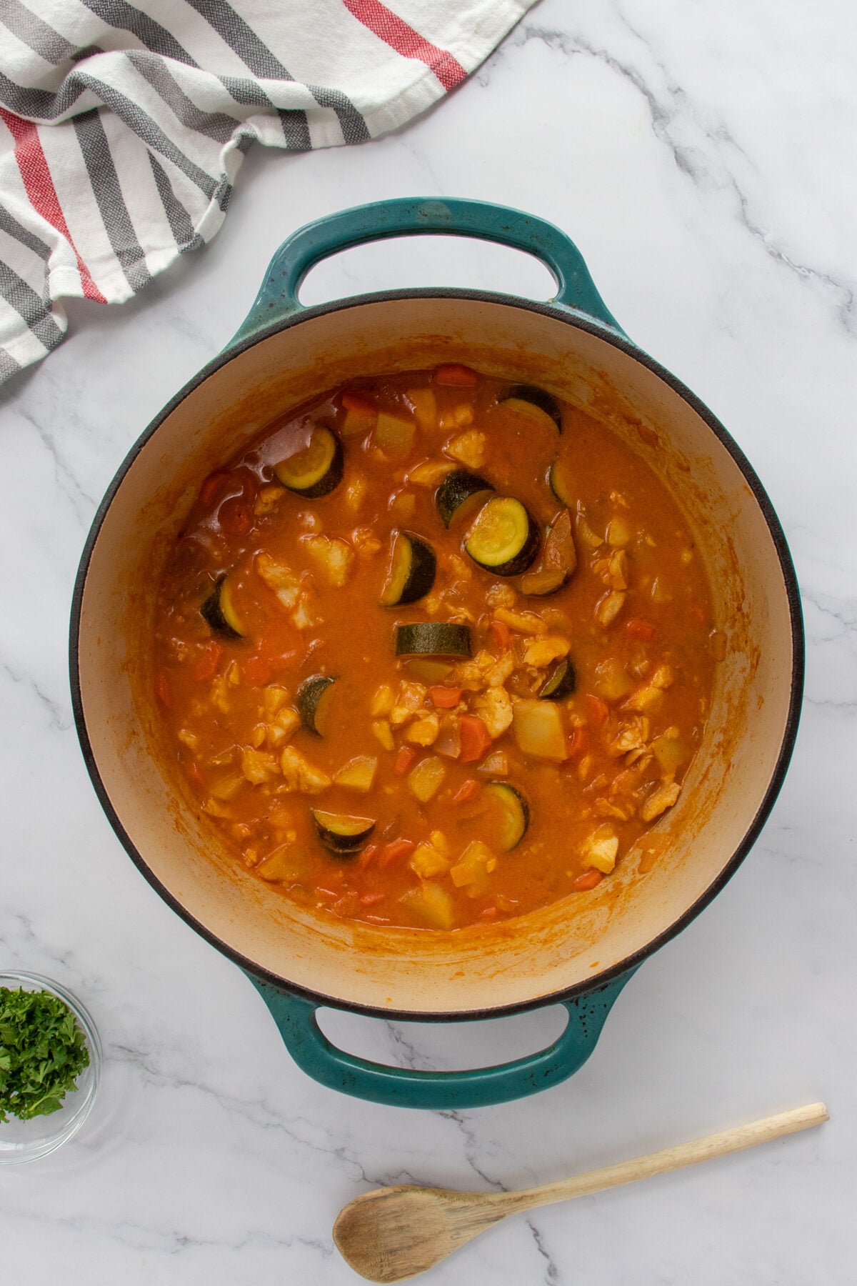 A teal Dutch oven filled with chunky vegetable stew sits on a marble countertop; a wooden spoon, a small bowl of chopped herbs, and a striped kitchen towel are nearby.