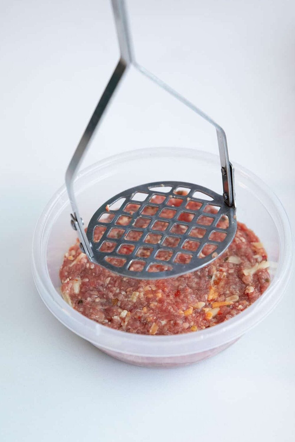 A metal potato masher presses down on raw ground meat mixed with diced onions and other ingredients in a clear plastic bowl against a white background.