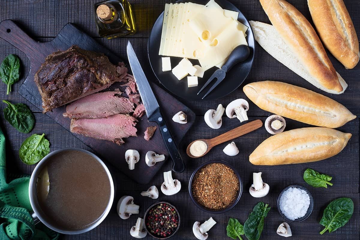 A top-down view of sliced roast beef, Swiss cheese, baguettes, mushrooms, spinach leaves, broth, spices, a knife, and olive oil arranged on a dark wooden table.