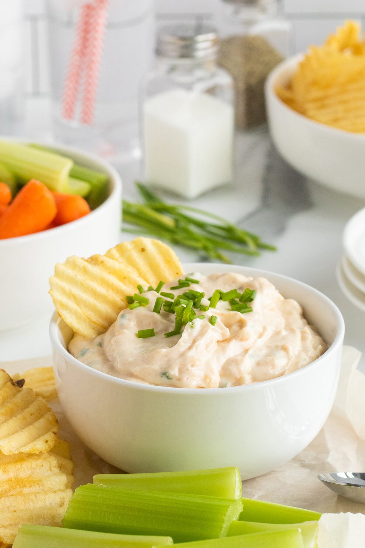 A bowl of creamy dip topped with chopped chives, surrounded by ridged potato chips and celery sticks, with more chips, vegetables, and seasonings visible in the background.