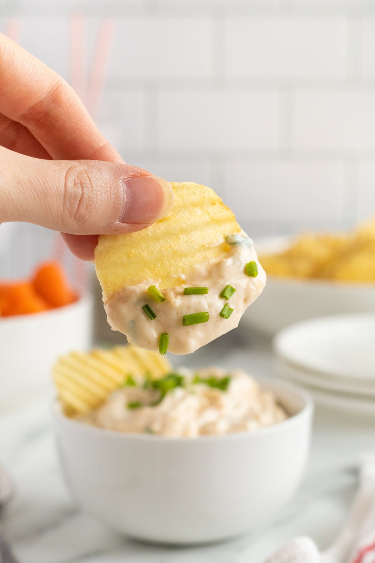 A hand holds a ridged potato chip dipped in creamy onion dip, topped with chopped chives. A bowl of dip with more chips and chives is visible in the background.
