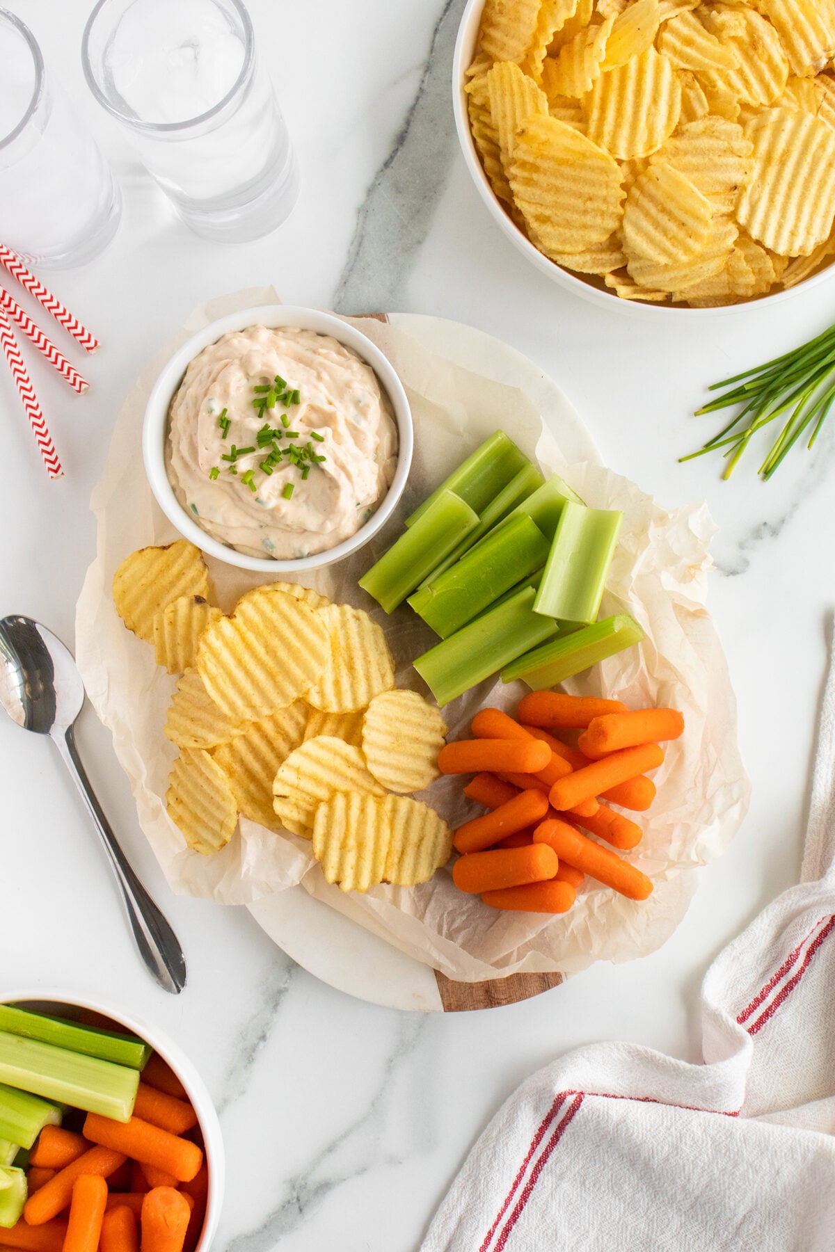 A white platter with ridged potato chips, baby carrots, celery sticks, and a bowl of dip garnished with chives. Nearby are bowls of chips, veggie sticks, glasses of water, and striped straws on a marble surface.
