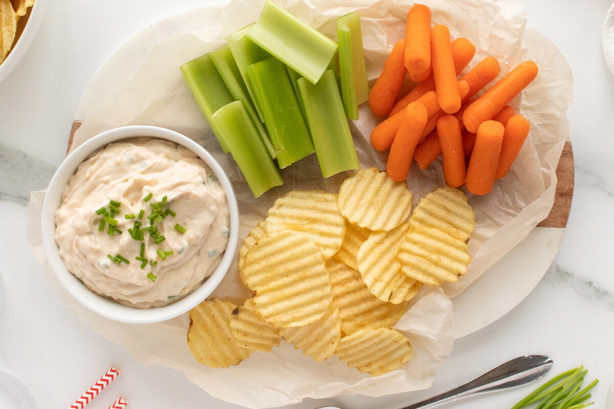 A platter with a bowl of creamy dip topped with chives, surrounded by celery sticks, baby carrots, and wavy potato chips, all arranged on parchment paper.