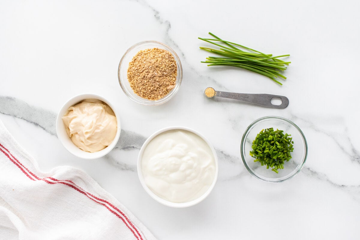An overhead view of small bowls with mayonnaise, breadcrumbs, and sour cream, a spoon with seasoning, chopped chives in a dish, whole chives, and a white towel with a red stripe on a white marble surface.
