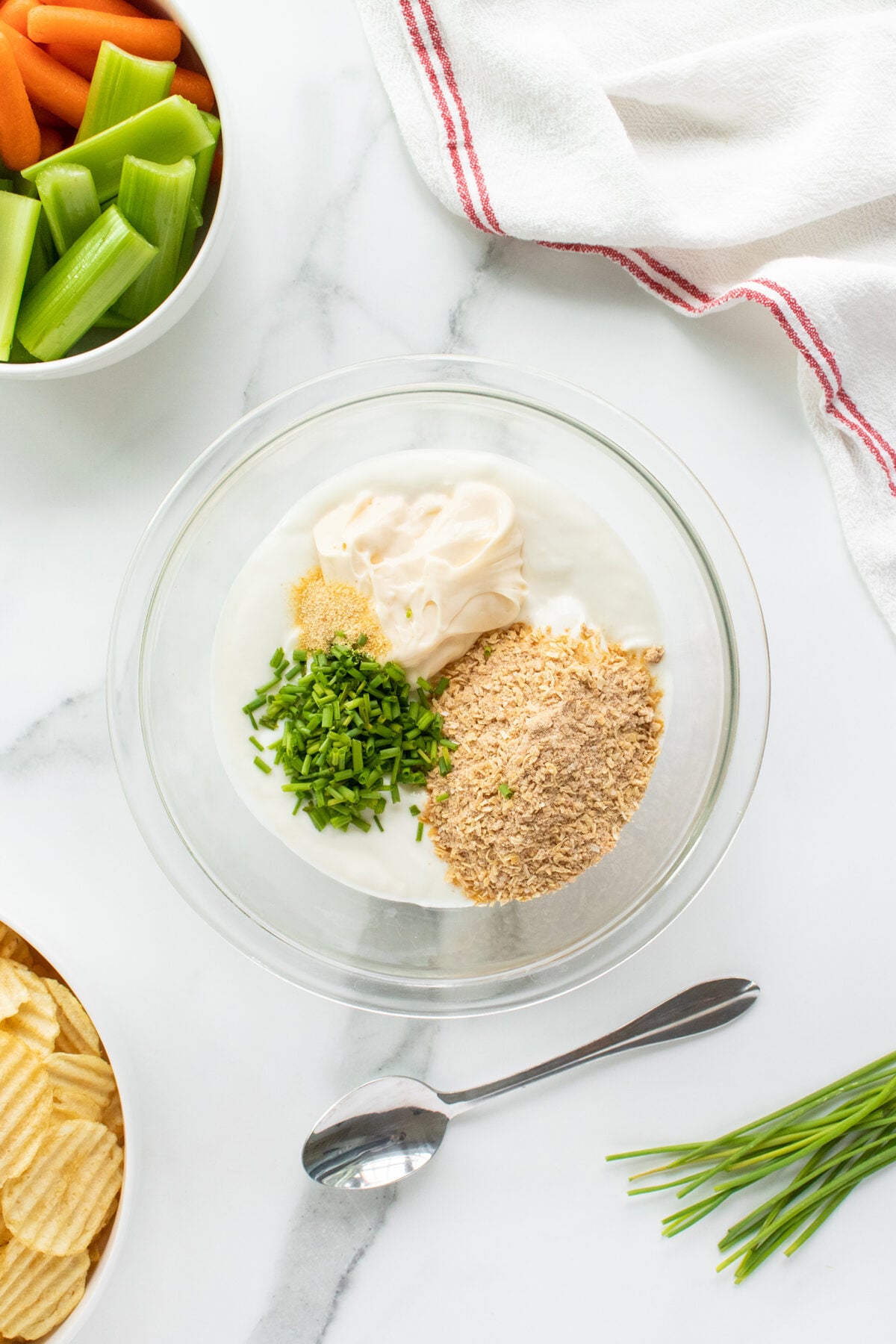 A clear glass bowl contains sour cream, mayonnaise, dried onion soup mix, chopped chives, and garlic powder on a marble countertop. Surrounding the bowl are potato chips, celery, carrots, a spoon, and a red-striped towel.