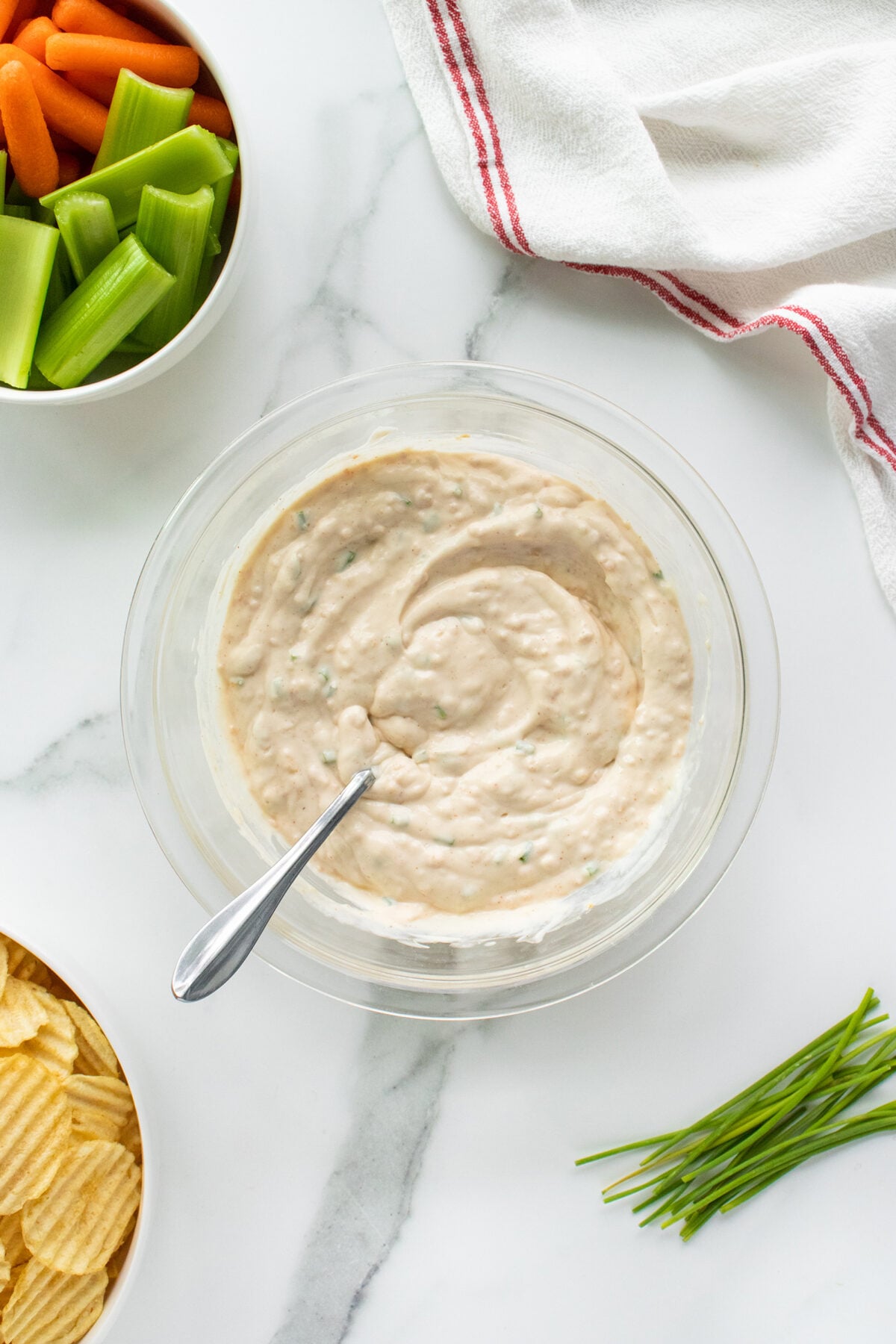 A bowl of creamy dip with a spoon sits on a marble surface, surrounded by a bowl of celery and carrots, a bowl of potato chips, fresh chives, and a white towel with a red stripe.