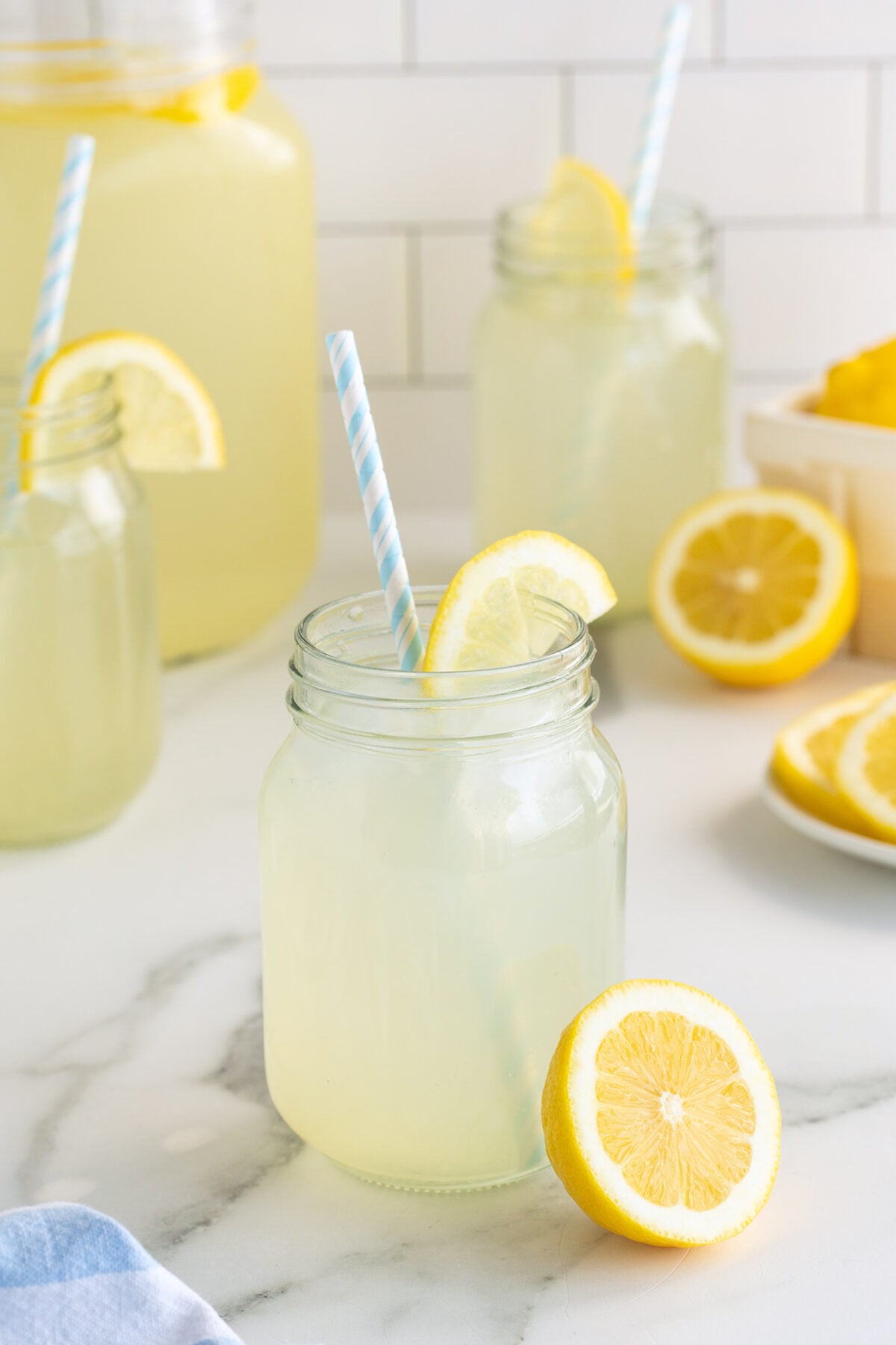 A glass jar of lemonade with a blue-striped straw and a lemon slice on the rim sits on a marble counter, surrounded by more jars of lemonade and cut lemons.