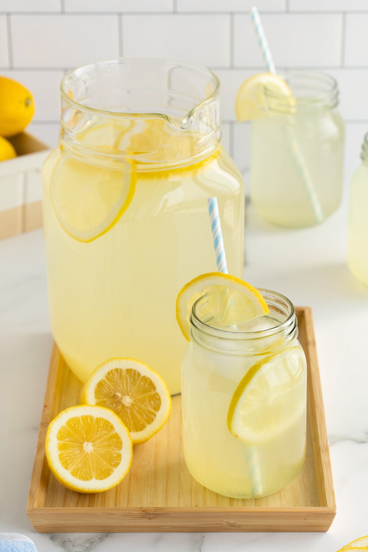 A pitcher and a mason jar of lemonade with lemon slices and striped straws sit on a wooden tray, with extra lemon slices nearby. Two more jars of lemonade are in the background on a white countertop.