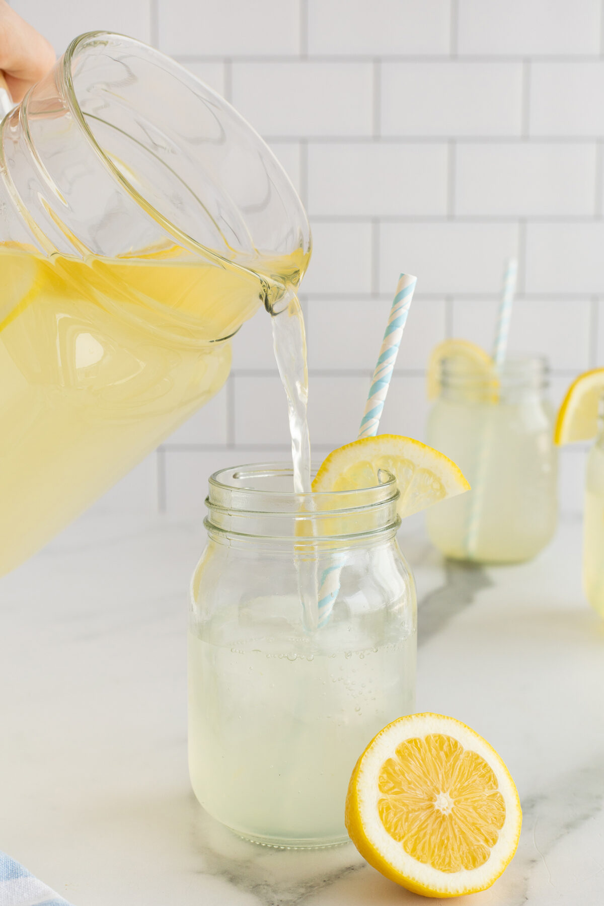 A pitcher of lemonade is being poured into a glass jar with a blue striped straw and a lemon slice. Two more jars of lemonade and lemon slices are in the background on a white surface with a tiled wall.