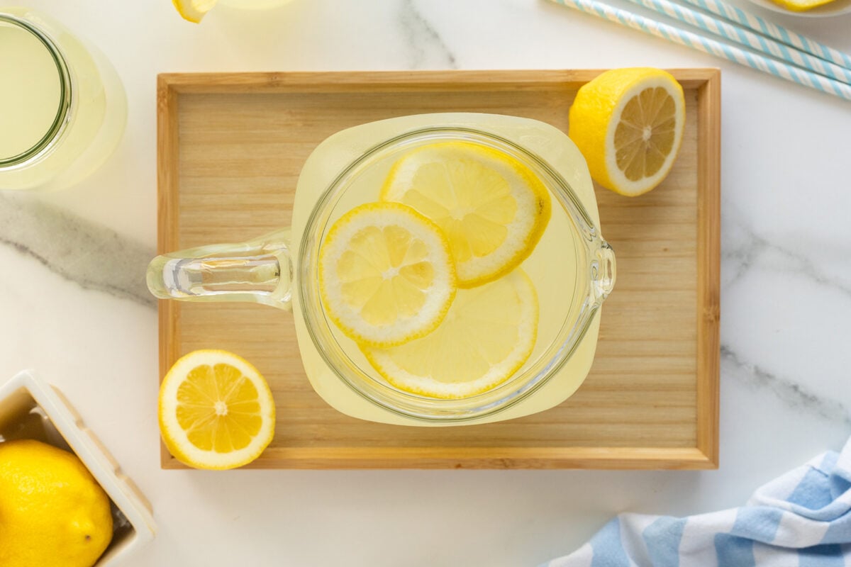 A glass pitcher filled with lemonade and lemon slices sits on a wooden tray, surrounded by lemon halves and a glass of lemonade on a white marble surface. Blue striped straws and a blue cloth are nearby.