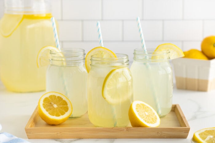 Three mason jars of lemonade with lemon slices and striped straws on a wooden tray. A jug of lemonade and whole lemons are in the background on a white countertop with a white tiled wall.